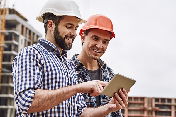 Working in team. Two young and cheerful builders in protective helmets are using digital tablet and working while standing at construction site